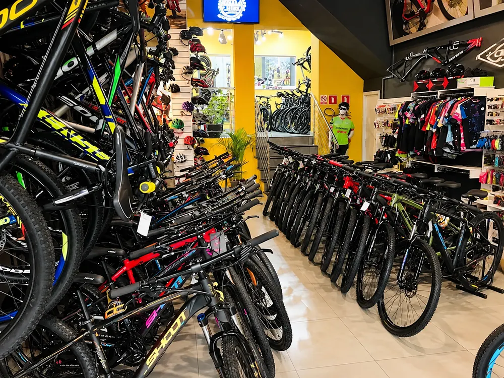 Salão da Bike Like com fileiras de bicicletas, parede de capacetes, escada amarela e araras de vestuário