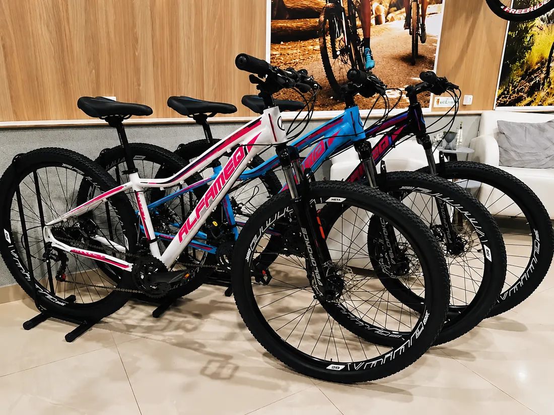Três mountain bikes nas cores branca e rosa, azul e preta, lado a lado em suportes na loja